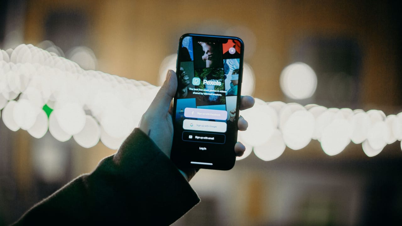 Mão segurando um celular com a tela de login de um aplicativo, diante de luzes desfocadas.