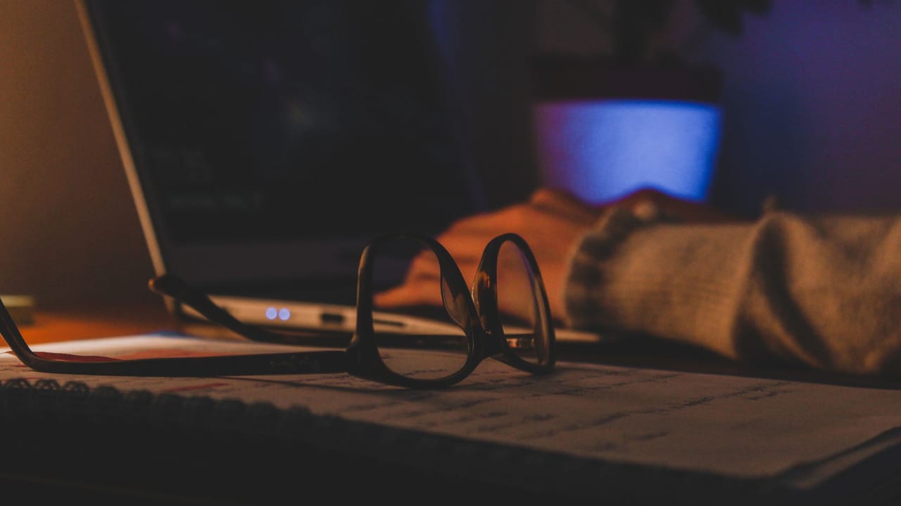 Óculos apoiados no teclado de um notebook aberto à noite, com a tela acesa ao fundo.