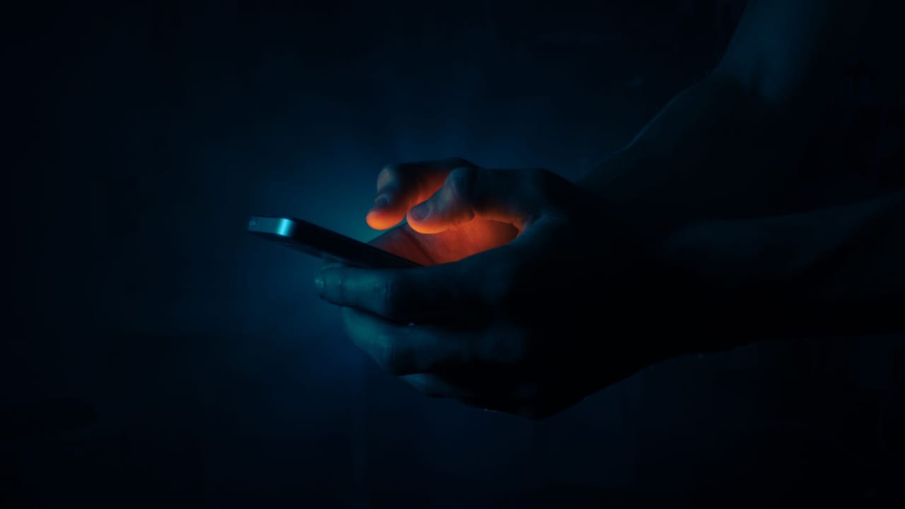 Mãos segurando um celular aceso no escuro, com o brilho azul da tela iluminando os dedos.