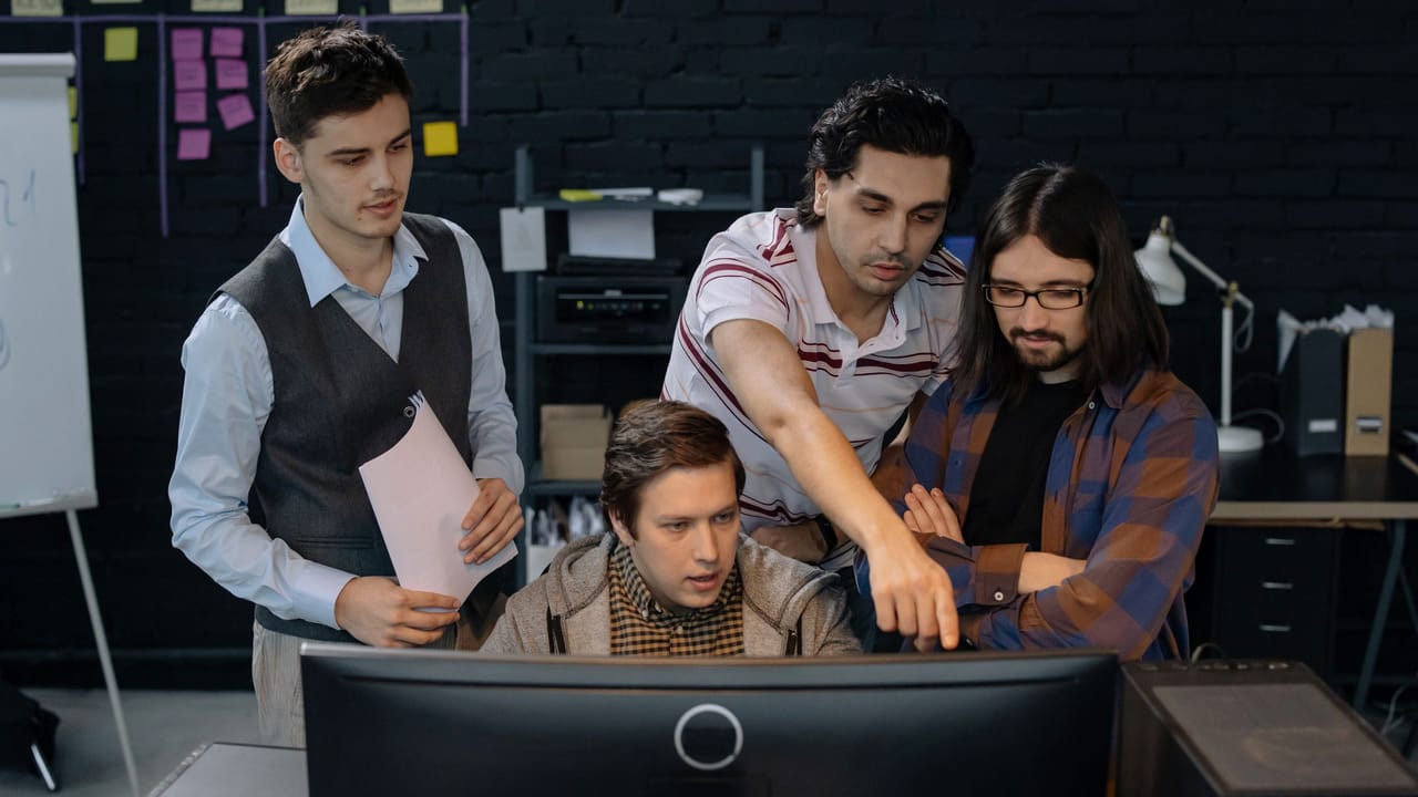 Equipe de desenvolvedores reunida em volta de um computador, discutindo um projeto num escritório.