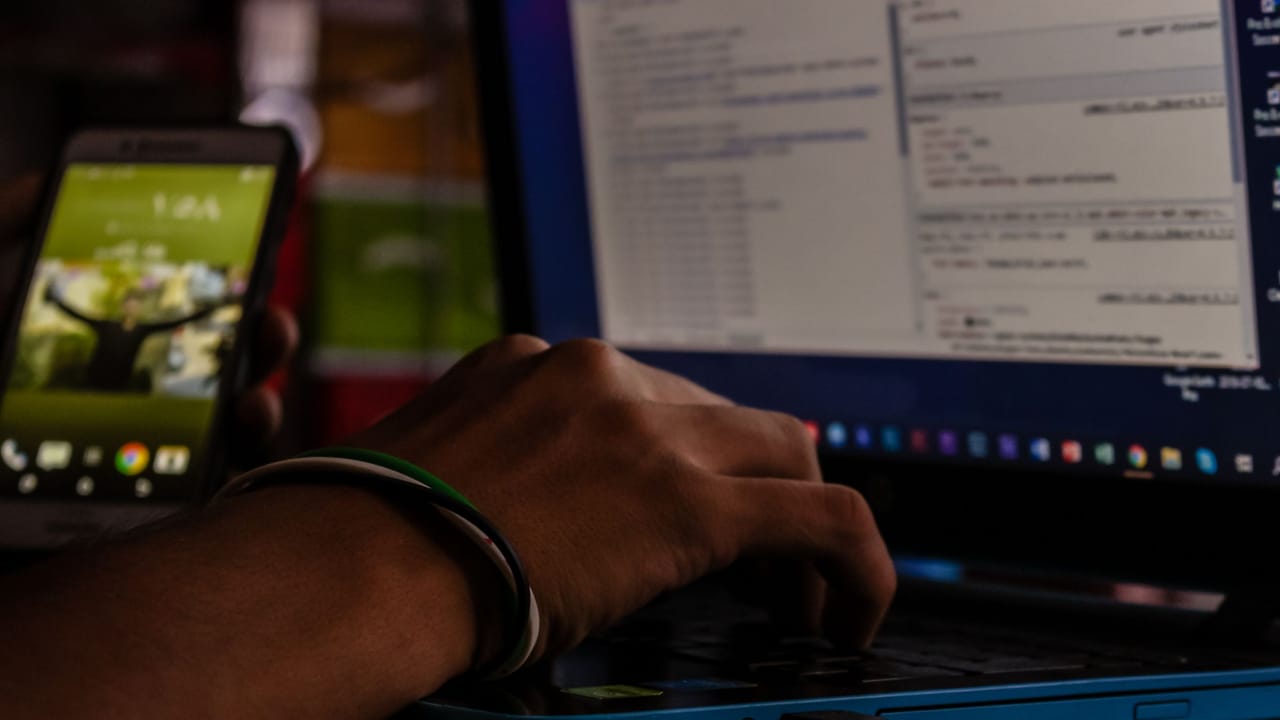 Mão digitando no notebook ao lado de um celular, num escritório pouco iluminado.