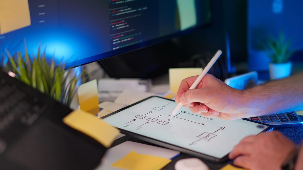 Mão desenhando um esquema num tablet, com código num monitor ao fundo e notas adesivas na mesa.