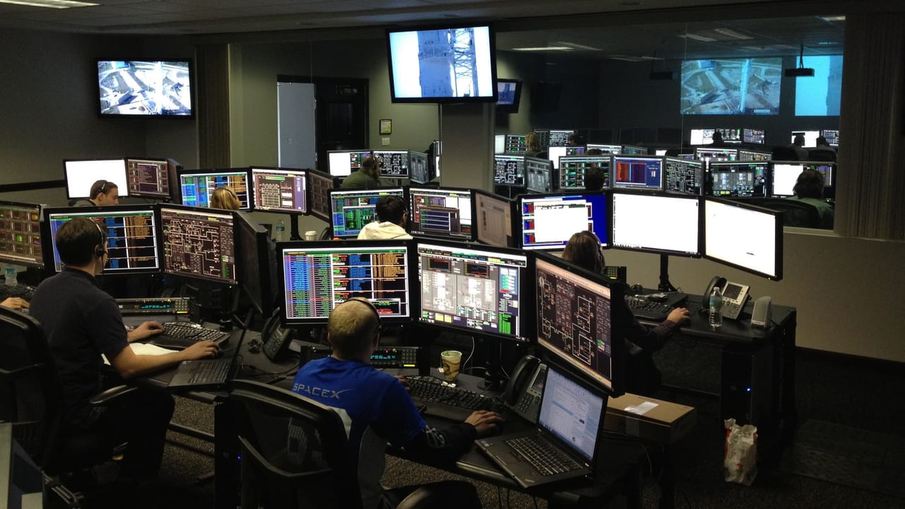 Sala de controle com pessoas trabalhando diante de vários monitores.