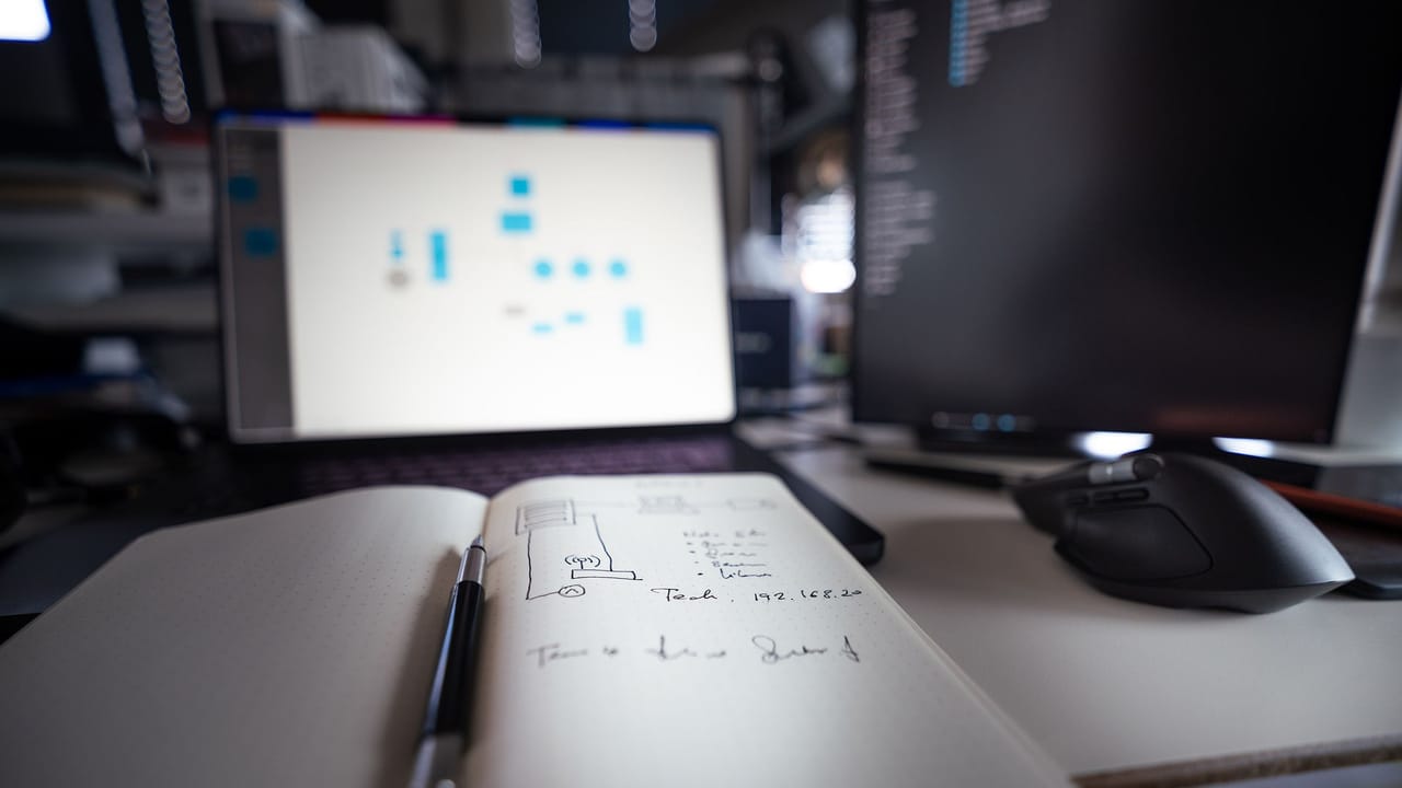 Caderno com o rascunho de uma página aberta sobre a mesa, diante de um monitor com o layout em construção.