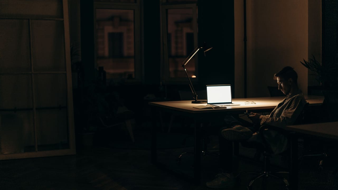 Homem trabalhando no notebook num escritório escuro à noite, iluminado por uma luminária de mesa.