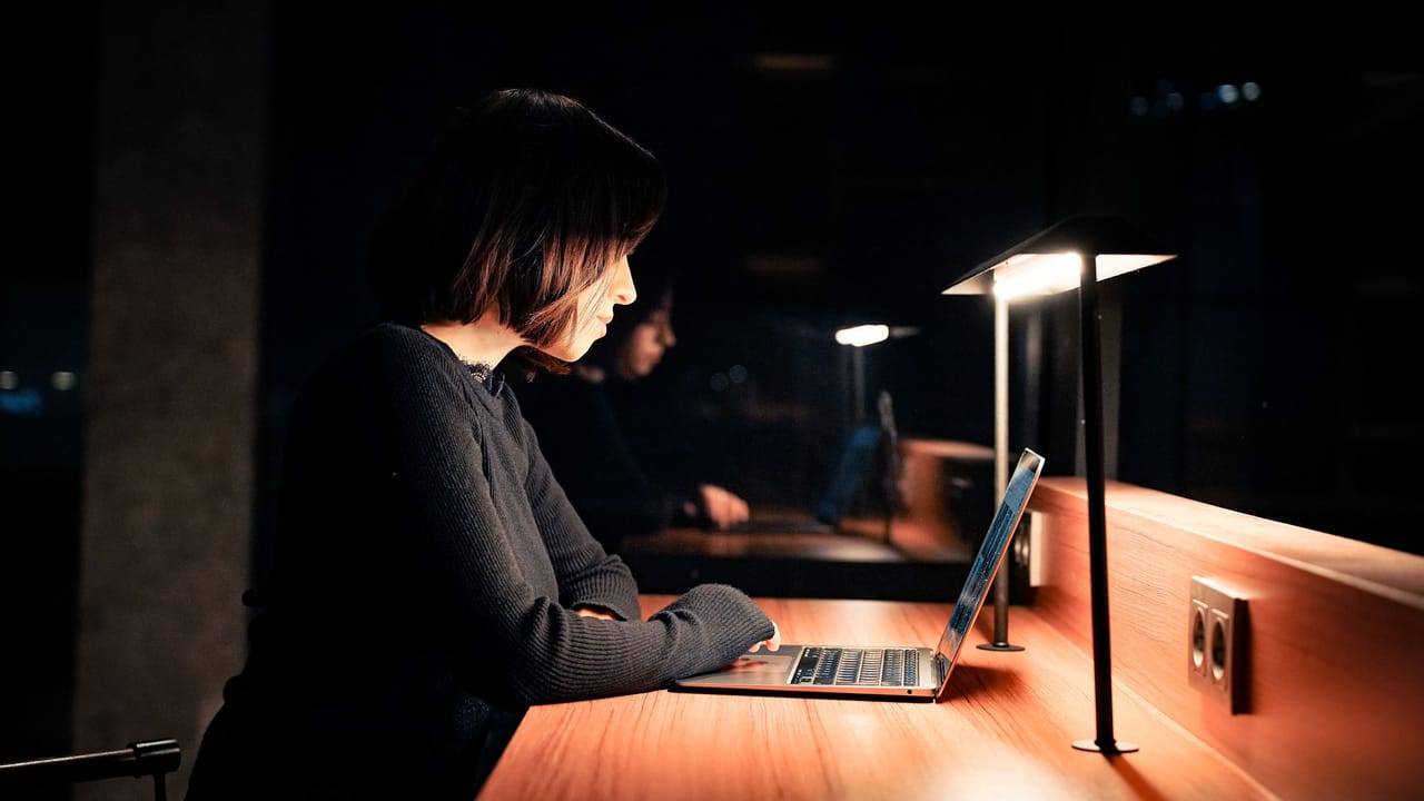 Mulher lendo no notebook à noite, sob a luz de uma luminária, numa mesa de escritório.